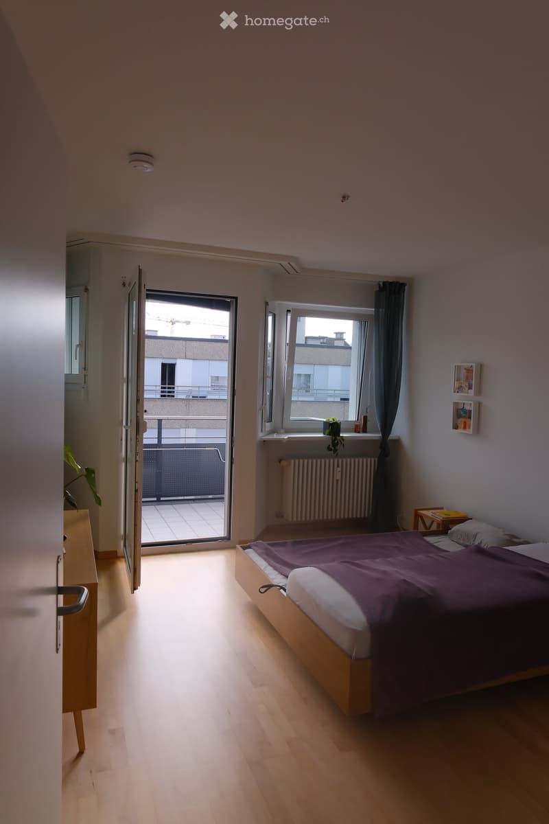 Bright bedroom with wooden flooring, a bed with purple bedding, a nightstand, a window with a curtain, and a sliding door leading to a balcony.