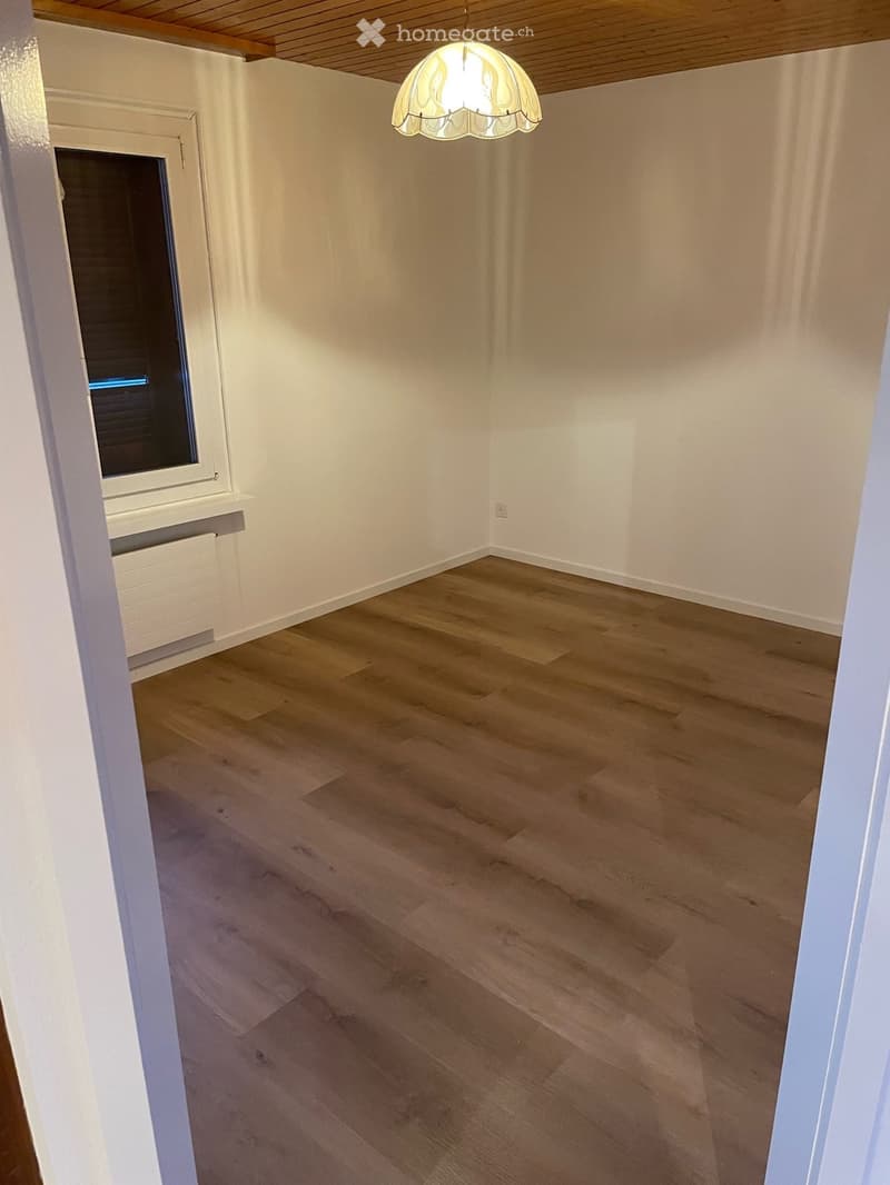 Empty room with wooden flooring, white walls, and a single window with blinds. A hanging light fixture is visible on the ceiling.