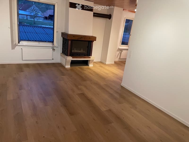 Empty room with wooden flooring, white walls, and a fireplace in the center. Two windows provide natural light, and radiators are visible on the walls.