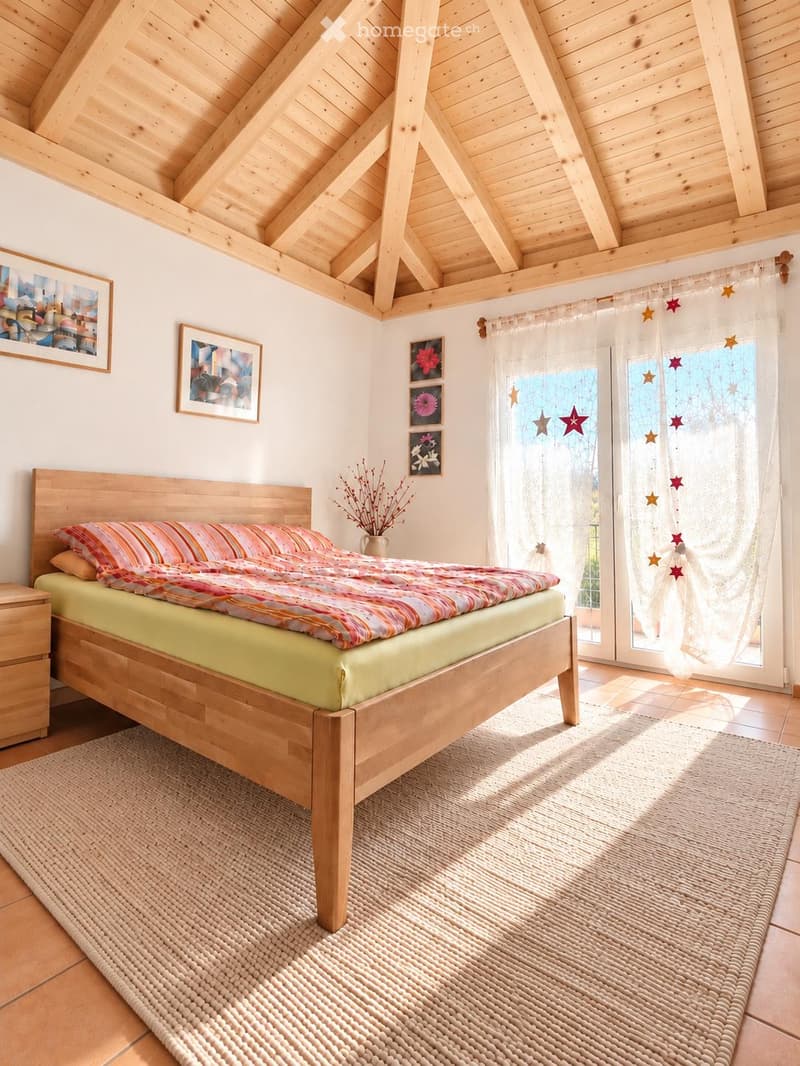 Cozy bedroom with wooden bed, colorful bedding, nightstand, woven rug, and decorative picture frames on the wall. Large window with sheer curtains allows natural light to fill the space.