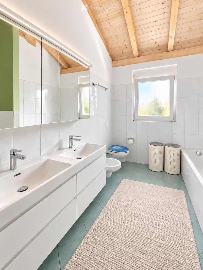 Modern bathroom with a double sink, toilet, bathtub, and large mirror. Features a light green tiled floor and white tiled walls, with a window providing natural light.