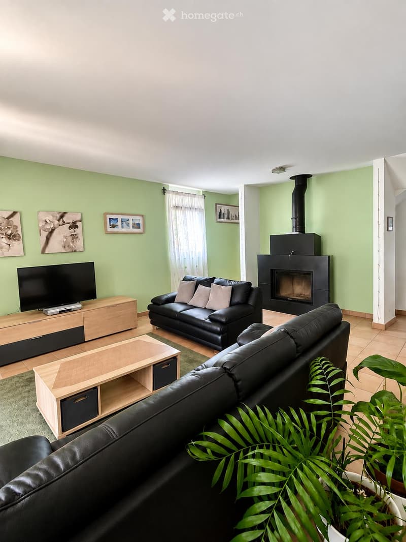 Bright living room with light green walls, black leather sofas, a wooden coffee table, a flat-screen TV on a wooden stand, and a fireplace with a black surround. A potted plant is in the foreground.