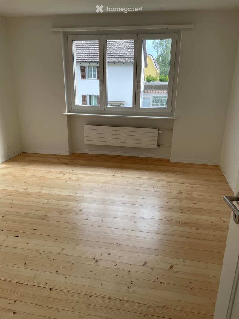 Empty room with light wooden flooring, white walls, and a large window with a radiator below it.