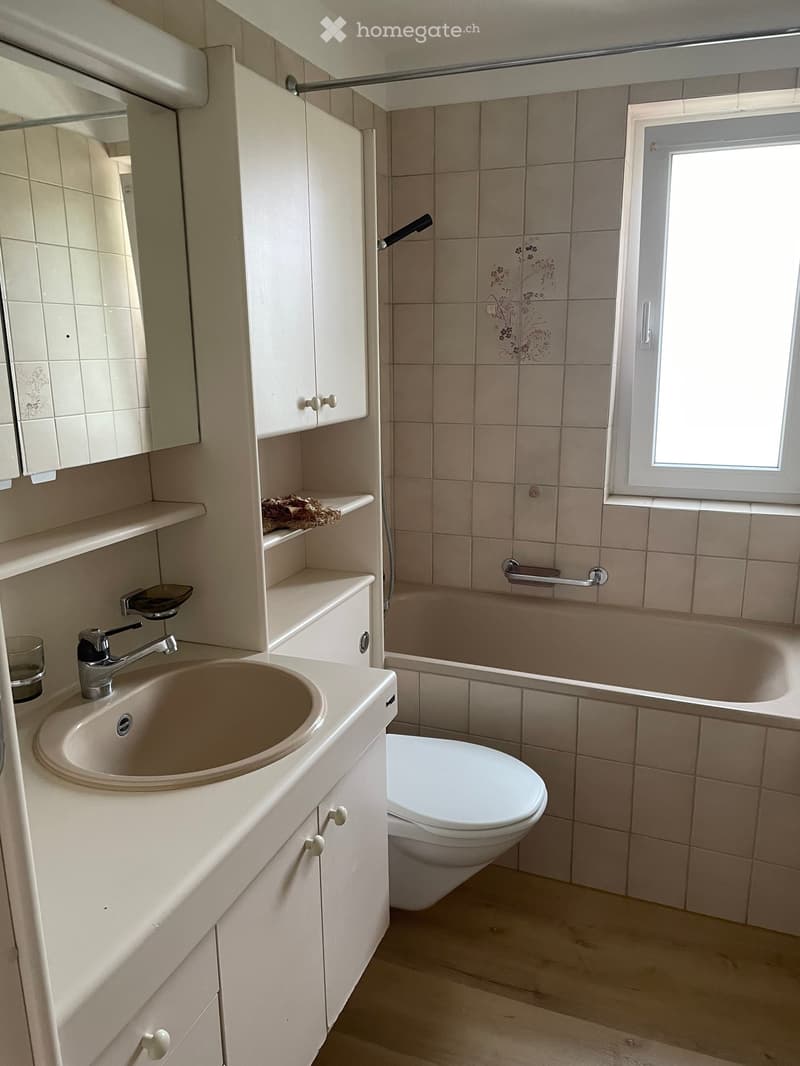 Bathroom with a sink, toilet, bathtub, and cabinet. Light-colored tiles and a window provide natural light.