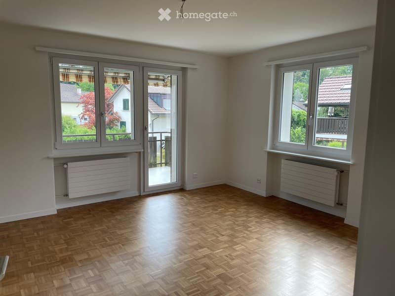 Empty room with wooden parquet flooring, large windows, radiators, and a sliding glass door leading to a balcony.