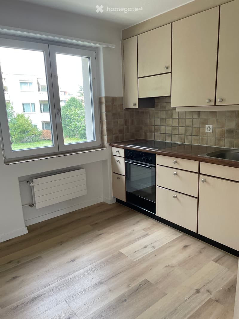 Modern kitchen with light wooden cabinets, a built-in oven, and a sink, featuring light wooden flooring and a large window with a view of greenery.