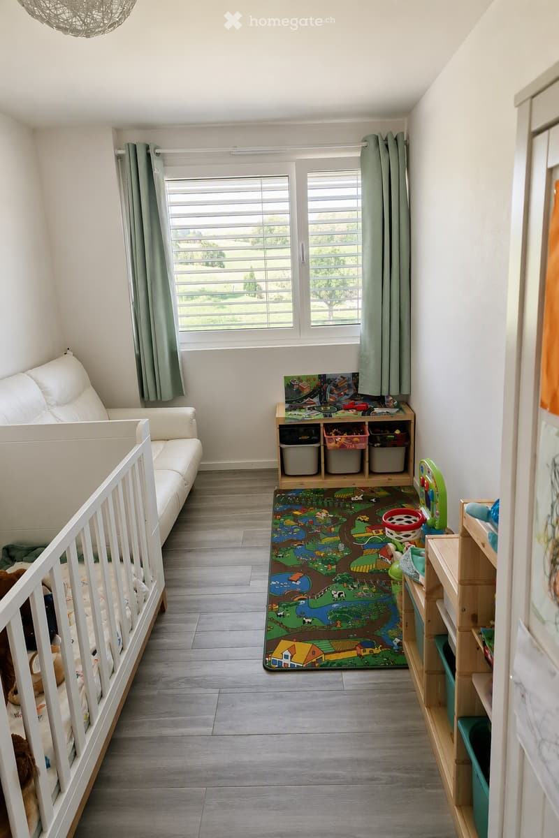 Bright, airy room with a white sofa, playpen, colorful play mat, storage unit, and shelves filled with toys. Large window with green curtains offers a view of greenery outside.