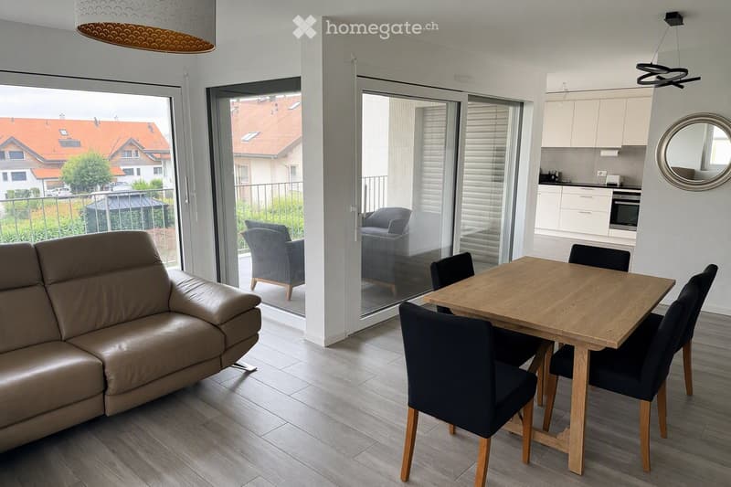 Modern living and dining area with a light-colored sofa, wooden dining table with chairs, and an open kitchen area with white cabinets and an oven. Large sliding glass doors lead to a balcony with outdoor seating.