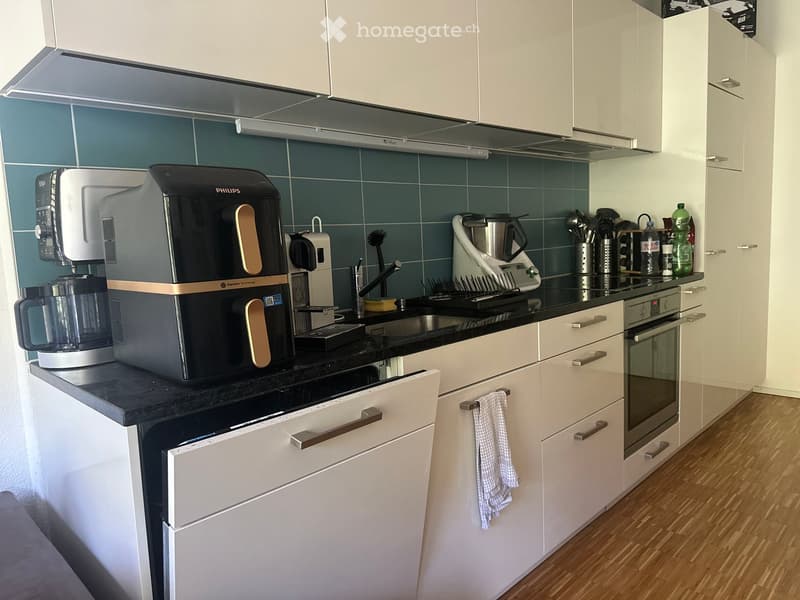 Modern kitchen with white cabinets, black countertops, and stainless steel appliances including a coffee maker, oven, and dishwasher. A towel hangs from a cabinet handle.