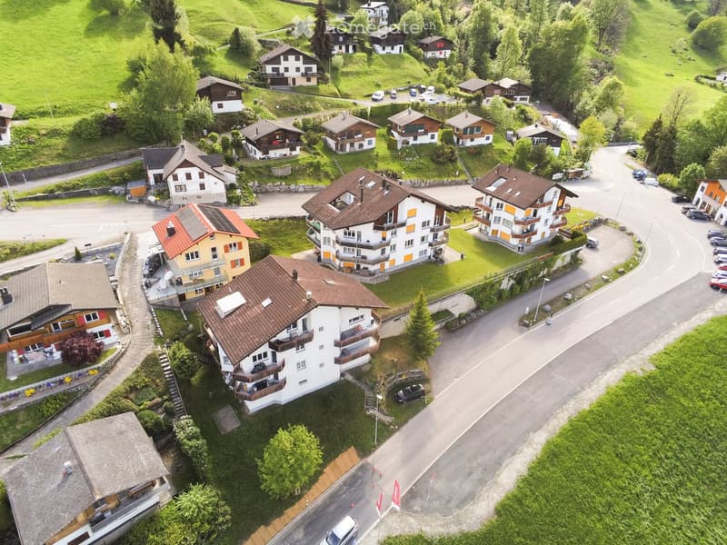 Aerial view of a residential area with multiple houses, surrounded by green hills and trees. Houses have varied architectural styles, with some featuring balconies and solar panels on the roofs. The area includes paved roads and parked cars.