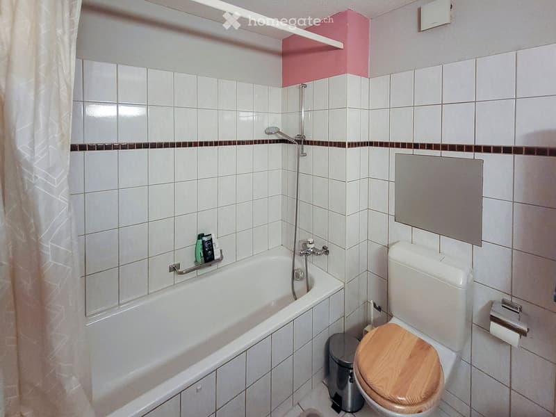 Bathroom with white tiled walls, a bathtub with a handheld shower, a toilet with a wooden seat, a mirror, and a small shelf holding toiletries.