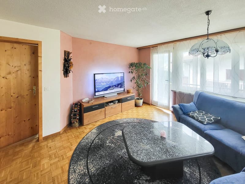 Living room with wooden flooring, a blue sofa, a glass coffee table, a television on a wooden stand, and a potted plant near the window.