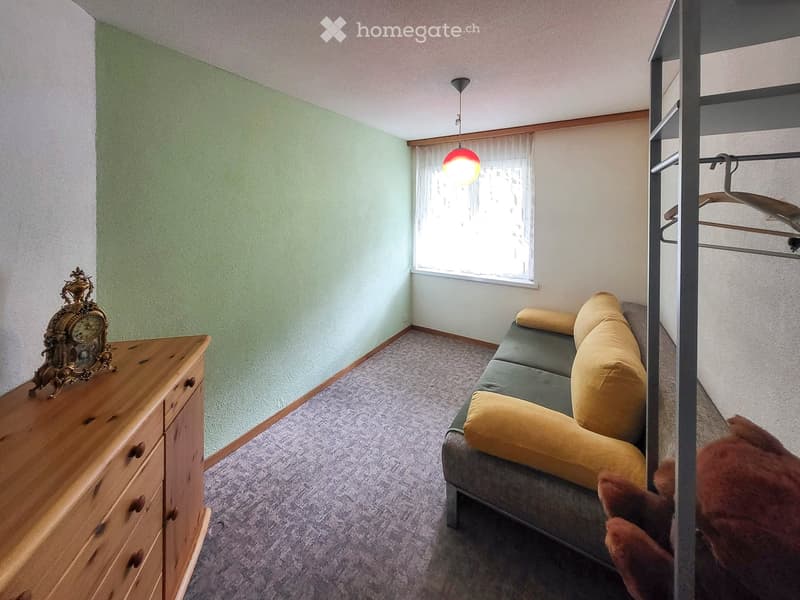 A furnished room with a grey sofa, yellow cushions, a wooden dresser, and a decorative clock on top. The room has light green walls, a window with white curtains, and a hanging light fixture.