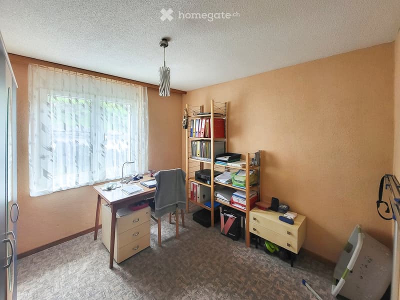 A furnished room with a desk, chair, shelf, and lamp. The room has a window with a curtain and a carpeted floor.