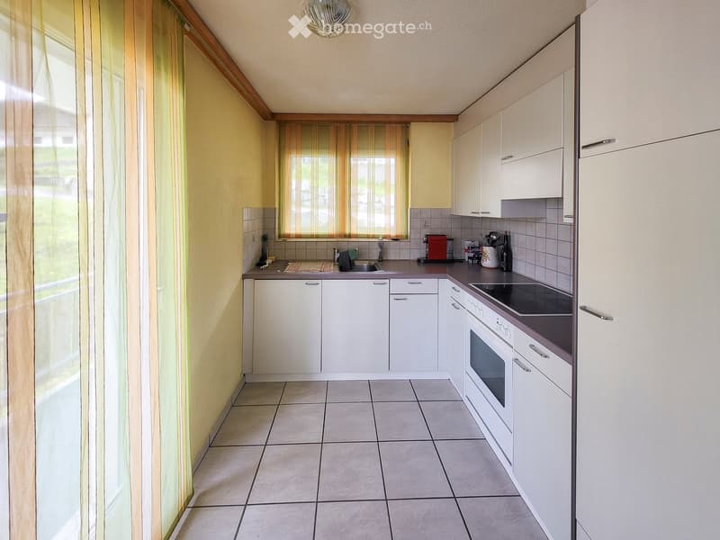 Modern kitchen with white cabinets, tiled floor, and stainless steel appliances including an oven, sink, and refrigerator. The space is well-lit with natural light from a window covered with striped curtains.