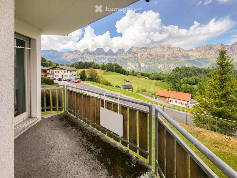 Balcony with metal railing and wooden fence, overlooking a scenic valley with mountains, houses, and green fields.