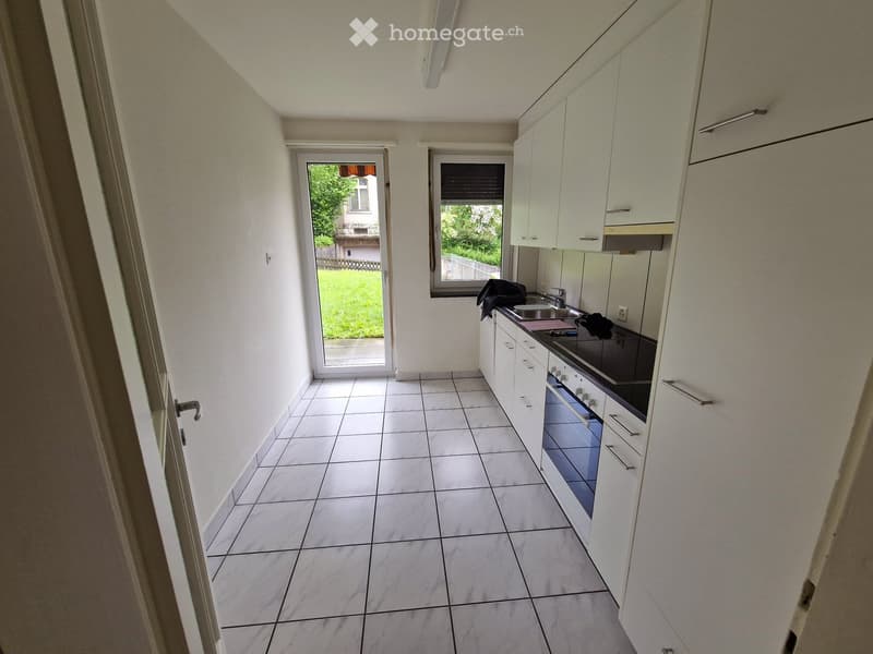 Modern kitchen with white cabinets, tiled floor, sink, and oven. Features a sliding glass door leading to a green outdoor area.