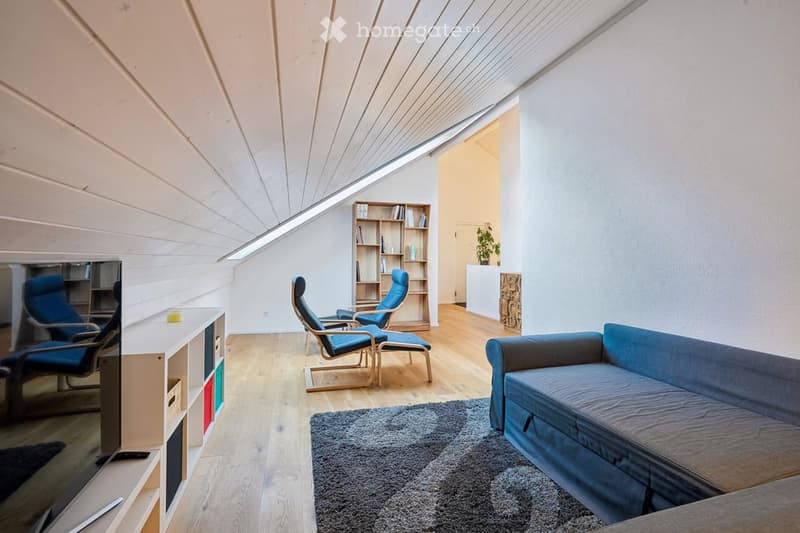 A cozy attic room with wooden floors, white walls, a blue couch, a bookshelf, and a TV. There are two blue chairs and a rug on the floor.