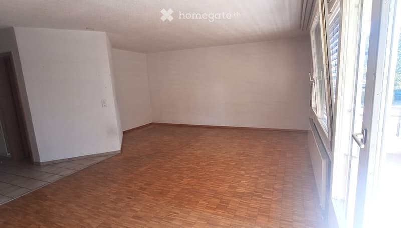 Empty room with wooden flooring and white walls, featuring a large window with blinds and a door leading to another area.