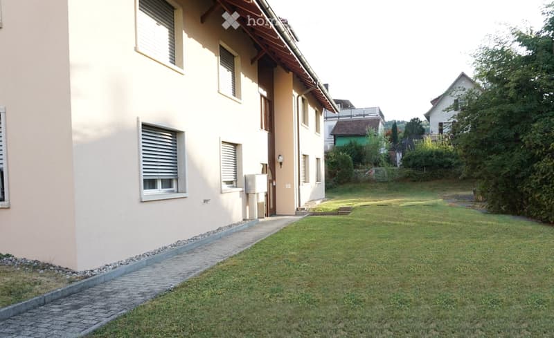 A well-maintained garden area with a paved pathway leading to a house entrance, surrounded by greenery and neighboring houses.