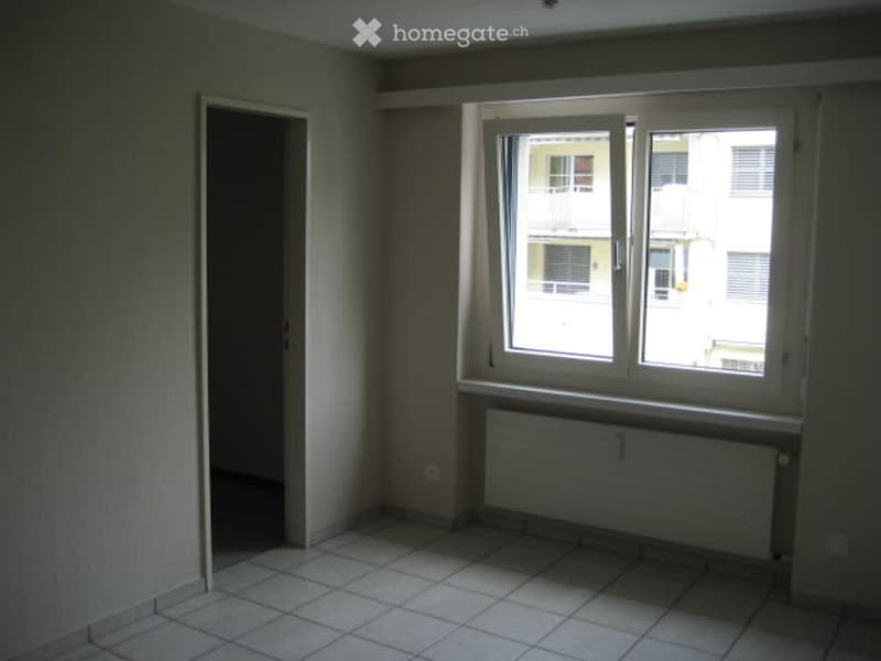 Empty room with tiled floor, white walls, a window with white frame, radiator under the window, and a doorway leading to another space.
