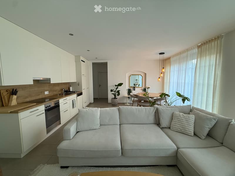 Modern, open-plan living and dining area with a grey sectional sofa, coffee table, kitchen with white cabinets, oven, sink, and dining table with chairs. Potted plants and a mirror are also present.