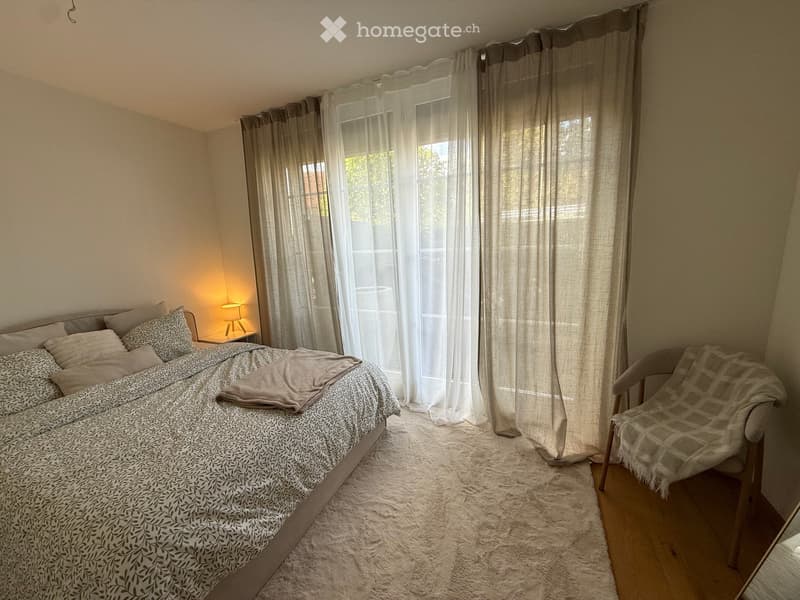 Cozy bedroom with a large bed, patterned bedding, a side table with a lamp, a chair with a throw, and sheer curtains covering a window.