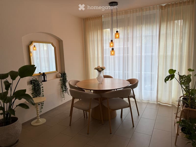 A modern dining area with a round wooden table, four chairs, a vase with white flowers, a large mirror, and potted plants.