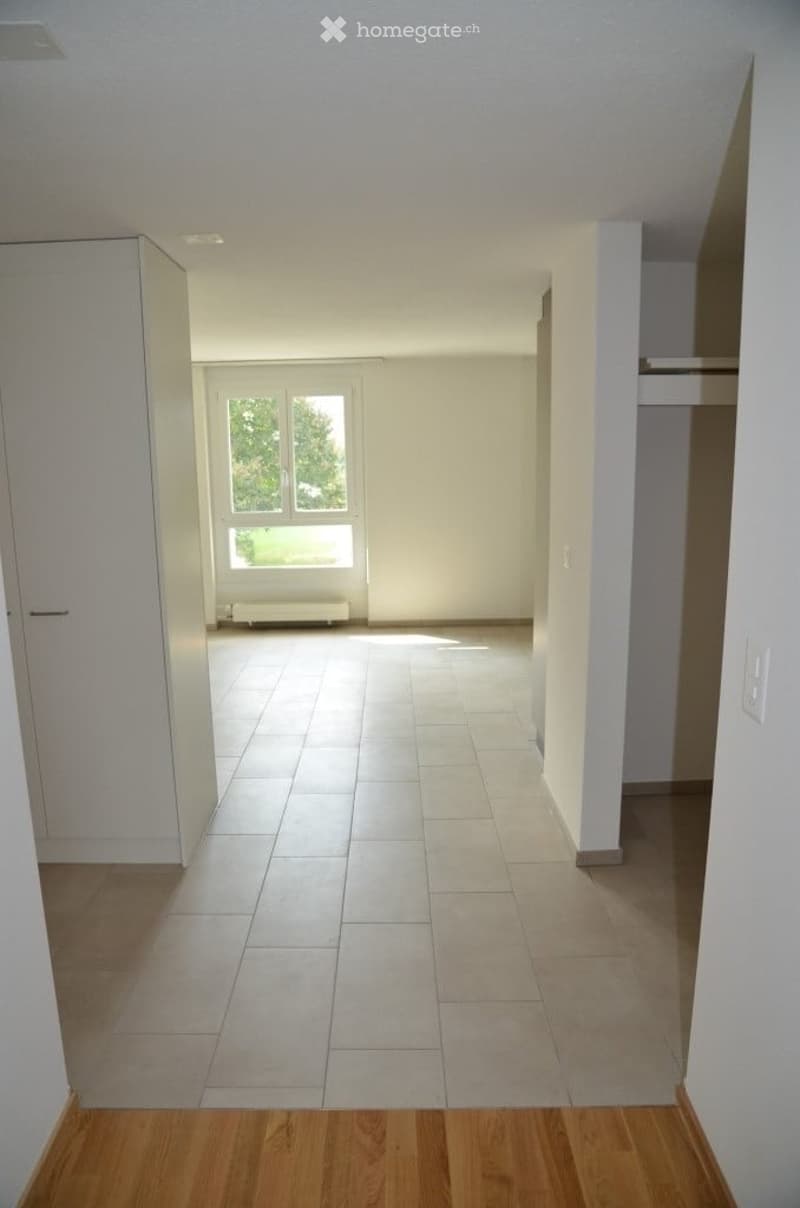 Empty hallway with tiled floor and white walls, leading to a room with large windows and a radiator.