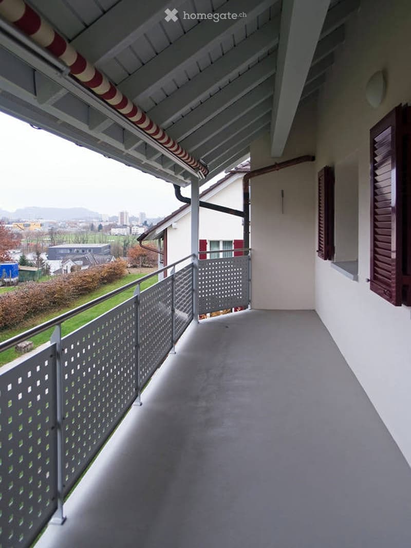 Balcony with metal railing, grey floor, and a view of the city and surrounding landscape.
