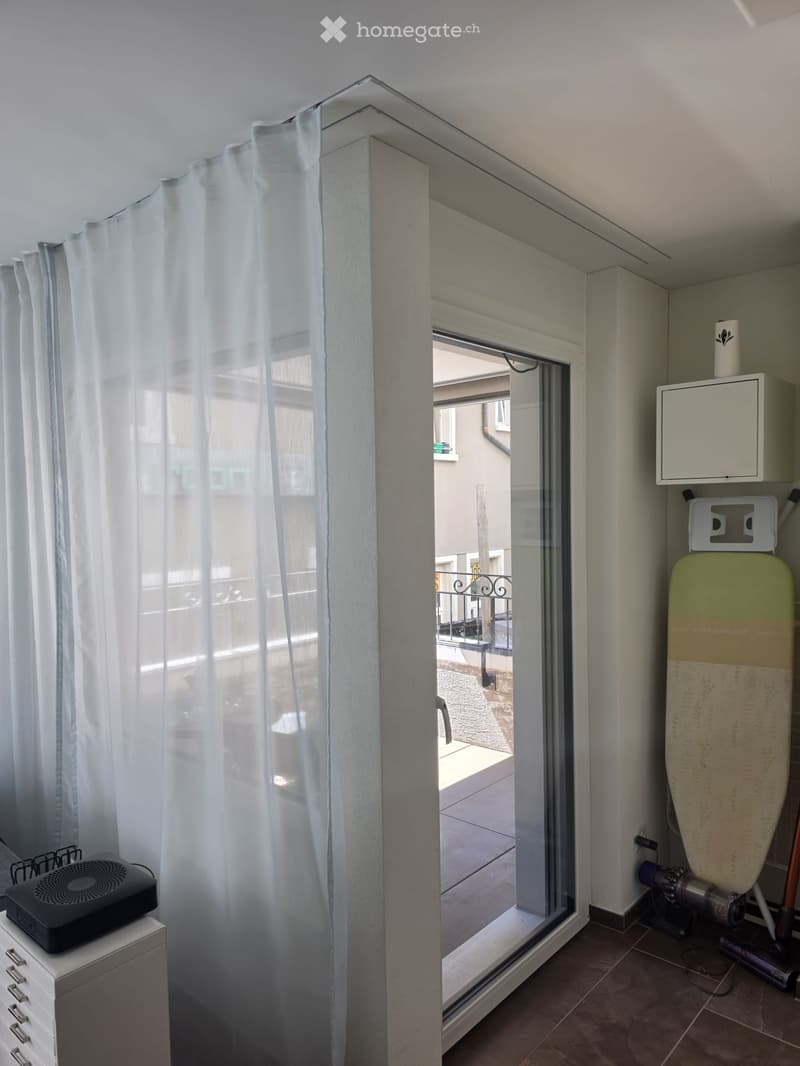 A small room with a large window leading to a balcony, featuring a curtain, an ironing board, a fan, and a small table with a drawer unit.
