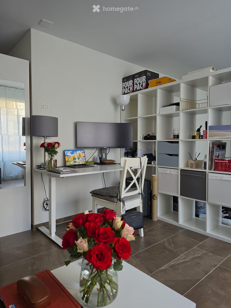 A modern, well-lit room with a white desk, laptop, screen, chair, and shelving unit. There is a vase with red and white flowers on a white table in the foreground.