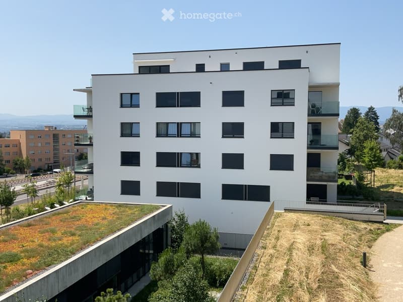 Modern multi-story residential building with white facade, large windows, balconies, and a green rooftop.