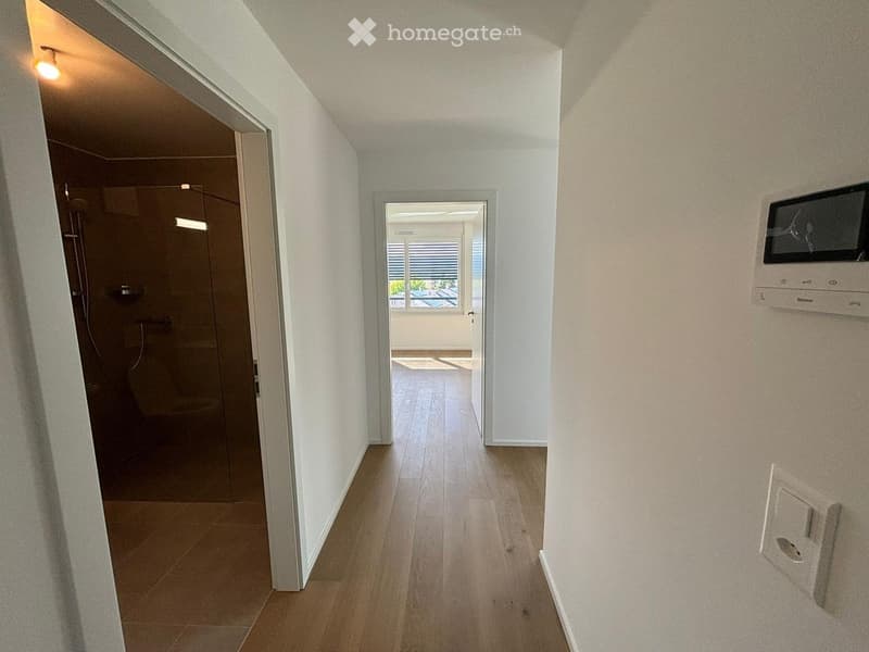 Empty hallway with wooden flooring, white walls, and doors leading to a bathroom and another room with a view.