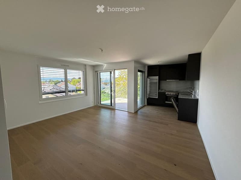 Open-plan unfurnished room with wooden flooring, large windows, and a sliding glass door leading to a balcony.