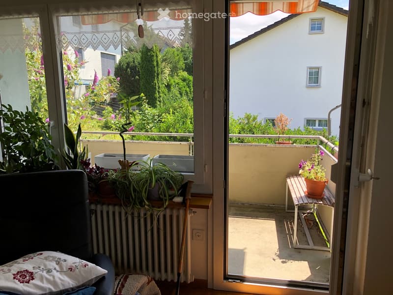 Balcony with wooden bench, potted plants, and view of greenery and neighboring building.