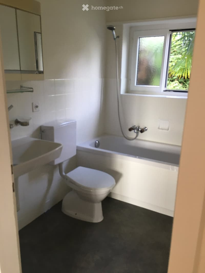 Modern bathroom with a toilet, bathtub, sink, mirror, and glass shelf. The room features white tiles and a window with a view of greenery.