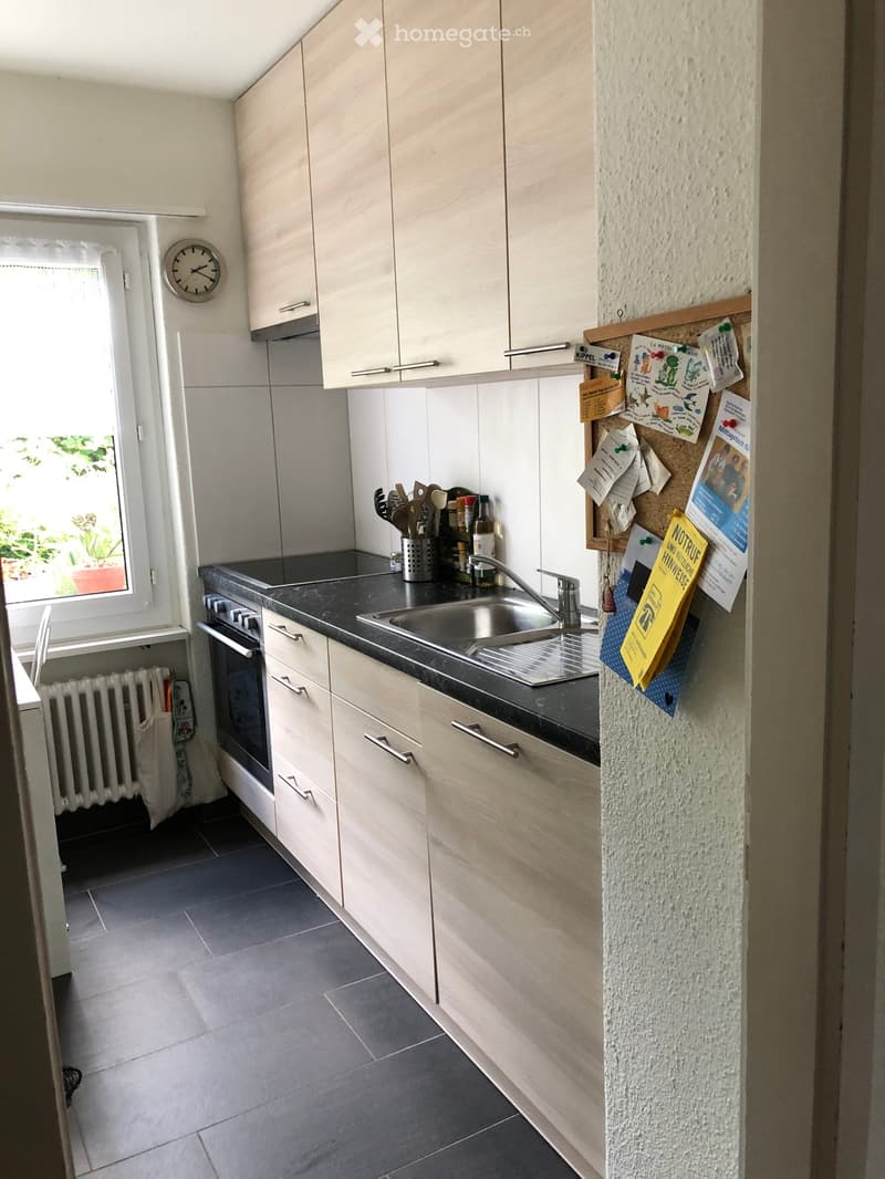 Modern kitchen with light wood cabinets, stainless steel sink, oven, and countertop. Features a window with blinds, a clock on the wall, and a radiator below the window.