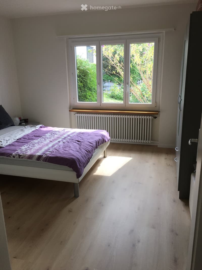 Bright bedroom with wooden flooring, a white bed with purple bedding, a radiator under a large window, and a nightstand with a lamp.