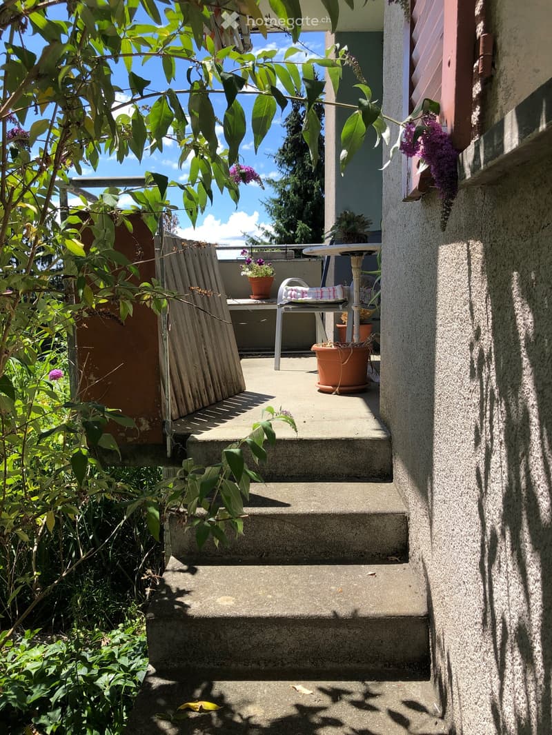 Balcony with a small table, chair, and flower pots, surrounded by greenery and steps leading up to it.