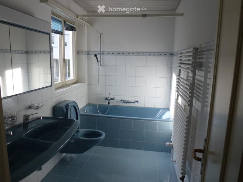 Bathroom with blue tiles, a sink, toilet, bathtub, and a showerhead. Features a window with blinds and a radiator.