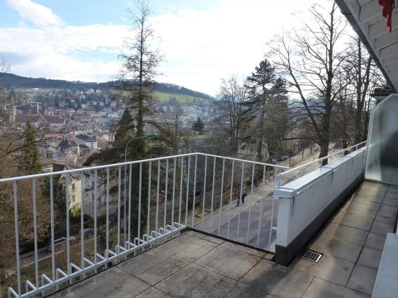 Balcony with white metal railing, tiled floor, overlooking a cityscape with trees and buildings.