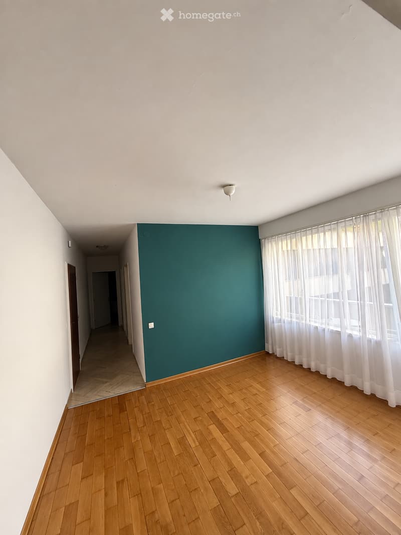 Empty room with wooden flooring, white walls, and a teal accent wall. Large window with white curtains allows natural light to enter. The room has a simple, modern design with no furniture or decorations.