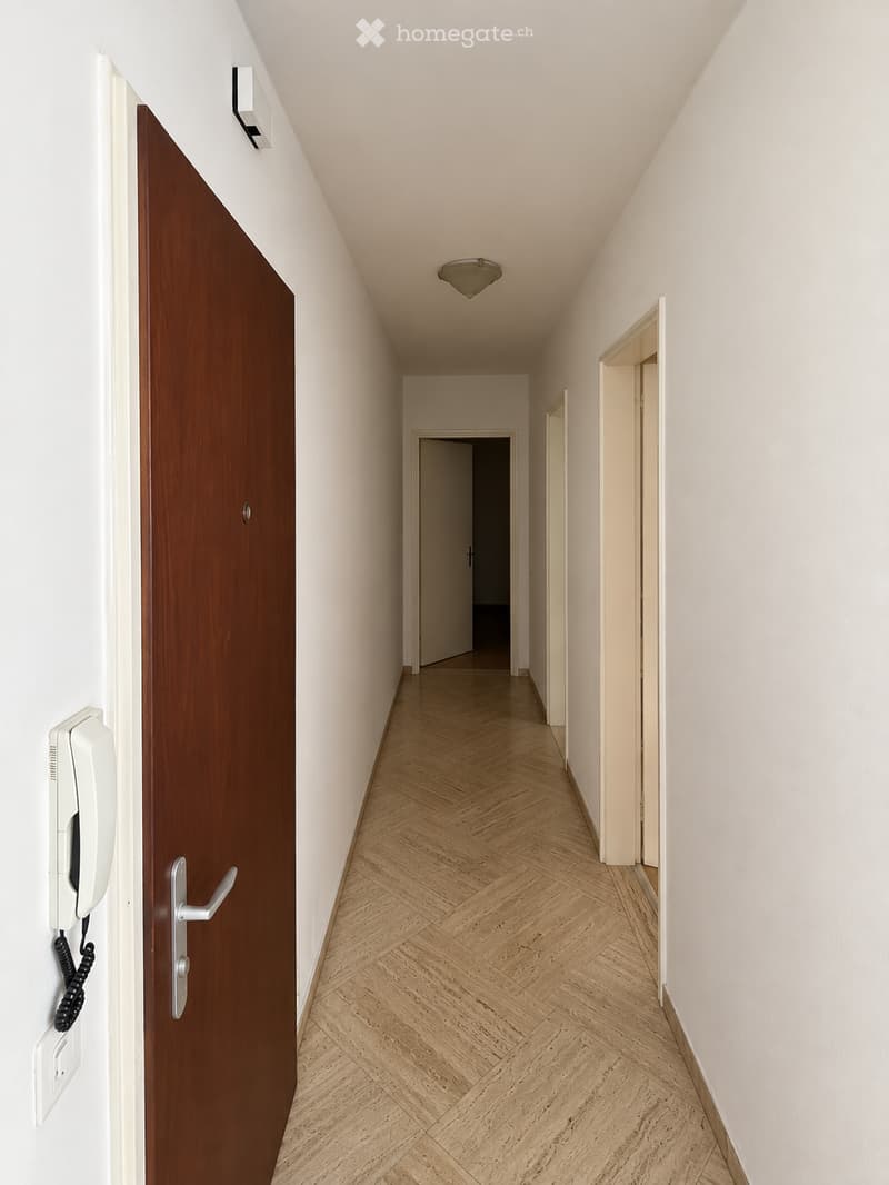 A narrow hallway with light-colored walls, wooden herringbone flooring, and a single ceiling light fixture. A closed wooden door is visible on the left, and an open doorway leads to another room at the end of the corridor.