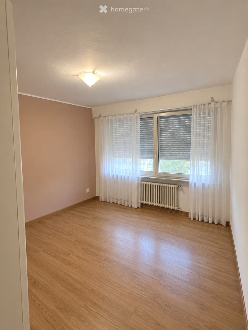 Empty room with wooden flooring, white walls, large window with white curtains, radiator, and a ceiling light fixture.