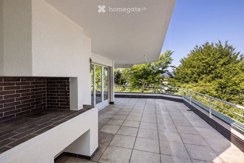 Spacious balcony with tiled flooring, white railings, and a view of lush greenery and trees.