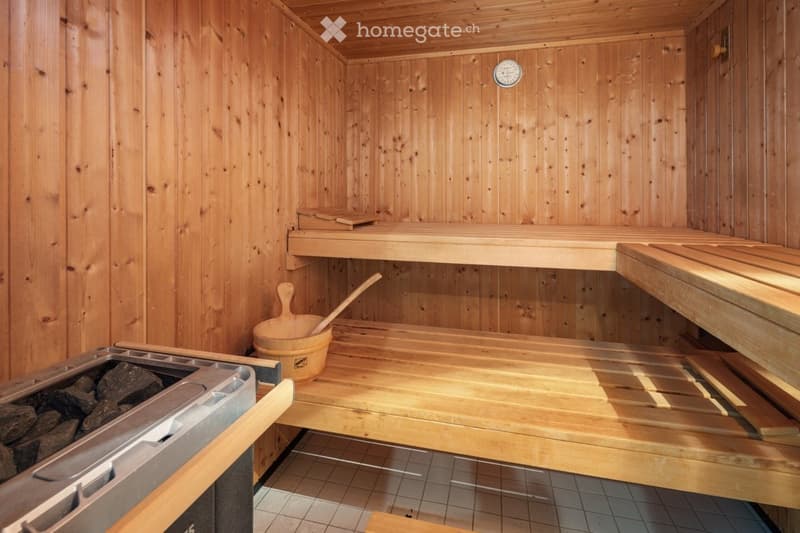 A wooden sauna with multiple levels of benches, a wooden bucket and paddle, a thermometer, and a stone bin for heating stones.