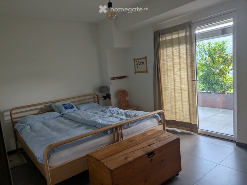 Cozy bedroom with a wooden bed, pillows, a wooden chest, a small table, and a chair. The room has light blue walls, a large window with curtains, and a view of greenery outside.
