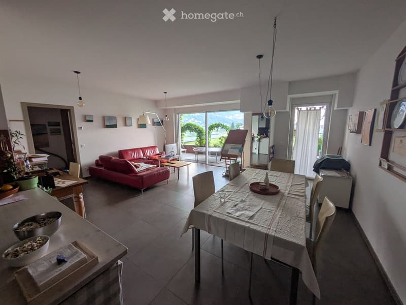 A well-lit living and dining area with a red leather sofa, a dining table with chairs, a coffee table, and decorative bowls on a side table. Large windows and sliding glass doors lead to a balcony with outdoor seating.