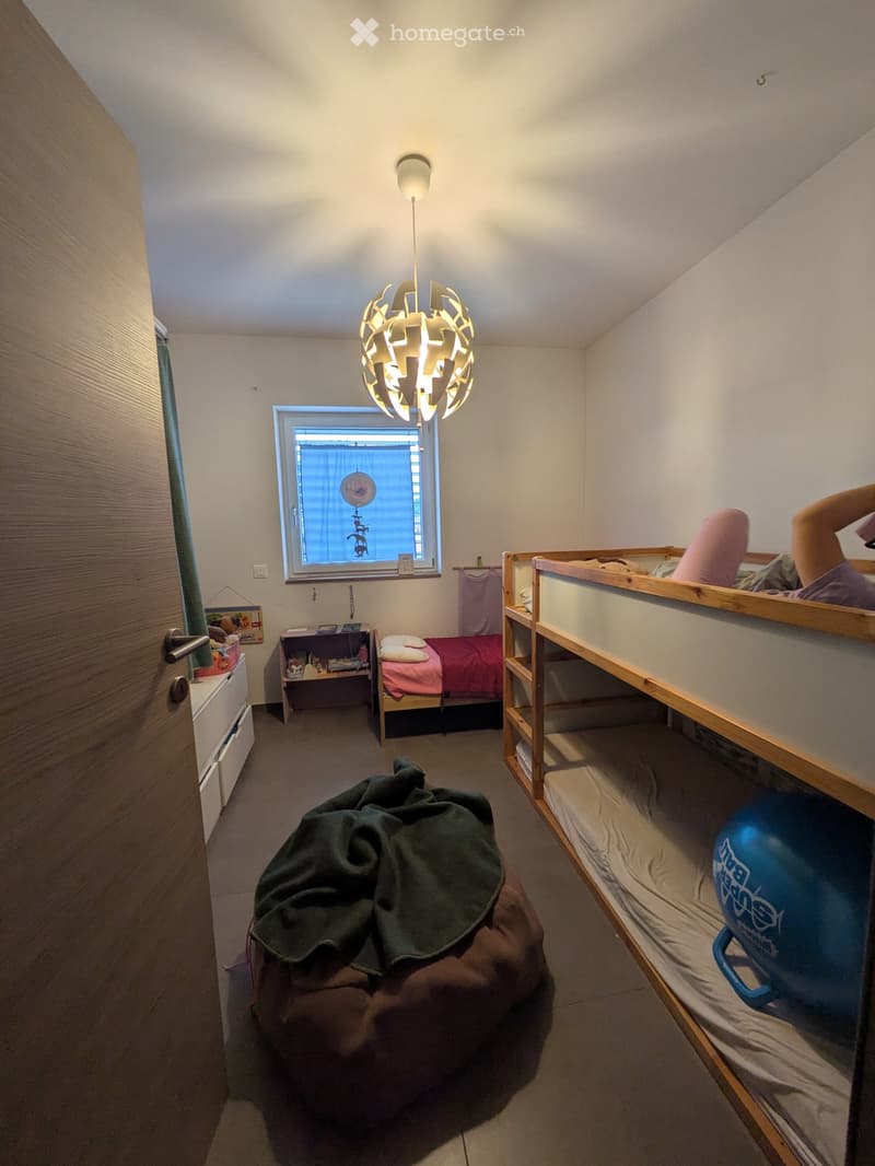 The room features a bunk bed, a single bed, a small table, a chair, and a lamp. There is also a green blanket on the floor and a blue ball on the lower bunk.